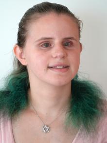 Lauren, a woman wearing a pink top and a Jewish Star necklace is smiling at the camera. Her hair, which is brown with streaks of turquoise at the bottom, is pulled back in a ponytail. 