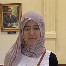 Amina, a young woman with light brown skin, stands inside the Texas Capitol on a decorative floor design. She wears a hijab, a white sleeveless sweater over a black long-sleeve shirt, wide-leg jeans, and black open-toed sandals. A small brown crossbody purse hangs at her hip, and she faces the camera with a neutral expression.