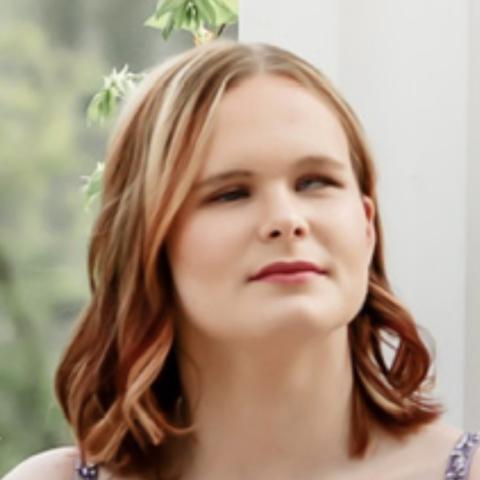Stormy, a young woman with auburn hair, smiles softly at the camera. She wears light makeup, and her hair falls loose around her shoulders.