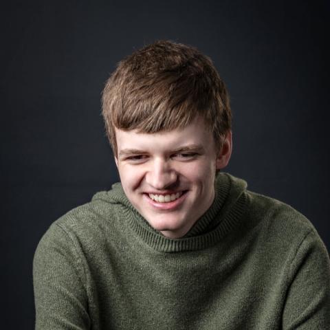 Benjamin, a young man with brown hair, sits against a gray background wearing a green shirt and smiling at the camera.